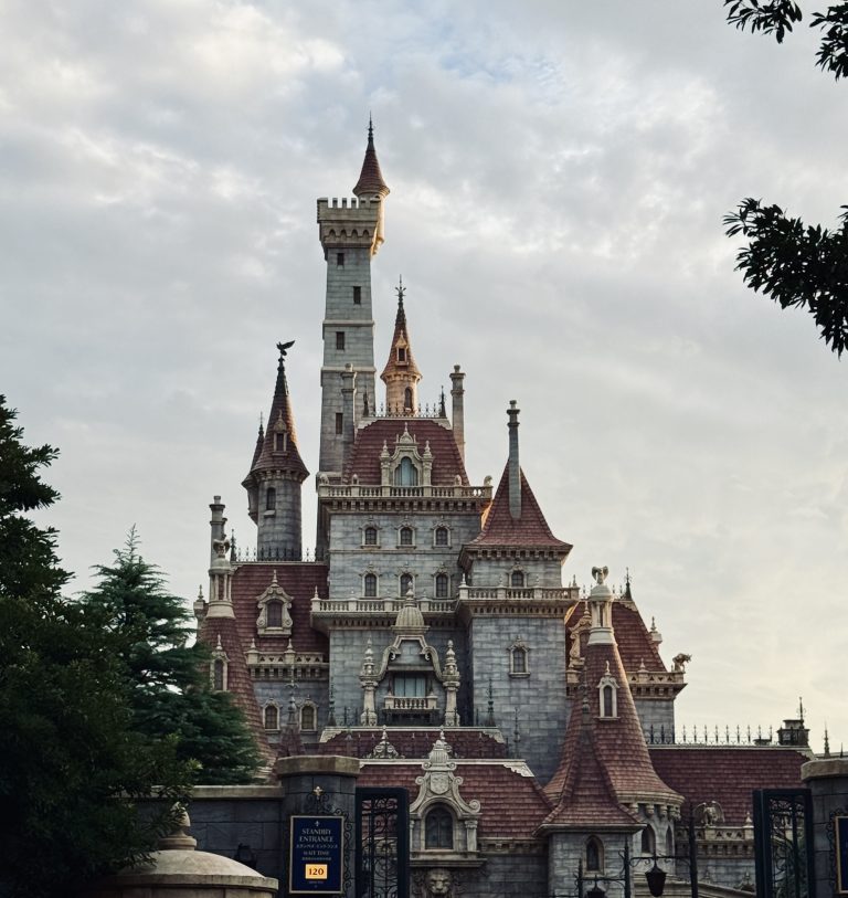 A grand, whimsical castle with multiple tall towers and intricate architectural details, featuring a mix of stone and red-tiled roofs. The castle is surrounded by lush greenery in the foreground under a cloudy sky.