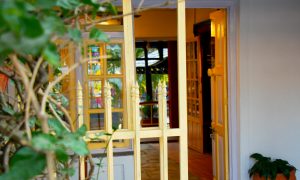 A view through an open yellow gate leading into a warm, welcoming space. Ornate wooden doors, glass panels, and green plants frame the entrance, while soft light inside creates a cozy atmosphere.