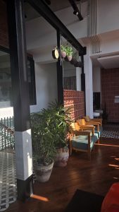 The image shows a modern interior space with a combination of wooden and tiled flooring. In the foreground, there are two wooden chairs with light blue cushions positioned near a potted plant. A decorative planter, resembling a face, holds small stones and greenery.