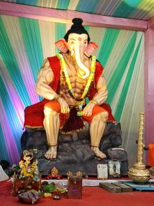 A statue of Ganesh in a tent. It's seated on a pile of rocks and there are offerings and other items at its feet. It's decorated for the Utsava celebration in Nikol, India.