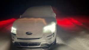 A snow covered car with its lights on in the dark.  The snow is lit white in the front and red in the back.
