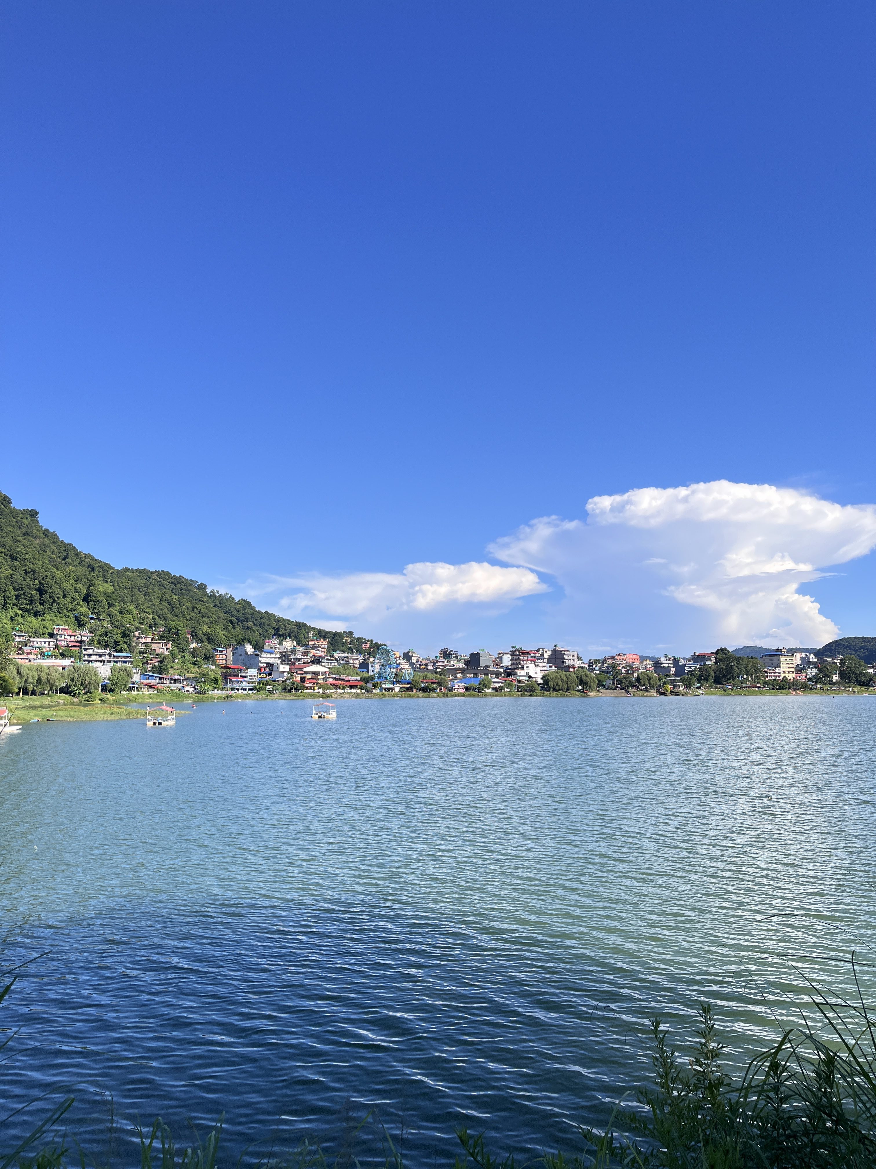 A serene lake surrounded by lush green hills under a clear blue sky. In the distance, colorful buildings line the shore, reflecting the vibrant colors of the houses in the water. 