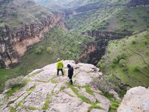 Two people stand on a cliff overlooking a green canyon with rocky mountains and steep slopes.