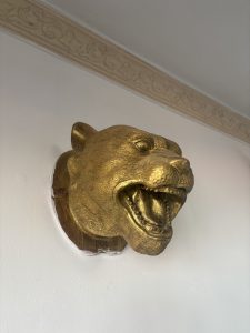 
A golden lion's head sculpture mounted on a white wall, displaying an open mouth and fierce expression, with detailed features such as eyes and ears