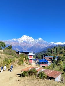 
A picturesque landscape featuring snow-capped mountains under a clear blue sky. In the foreground, a dirt path leads towards a small settlement with colorful buildings, including red, blue, and white rooftops