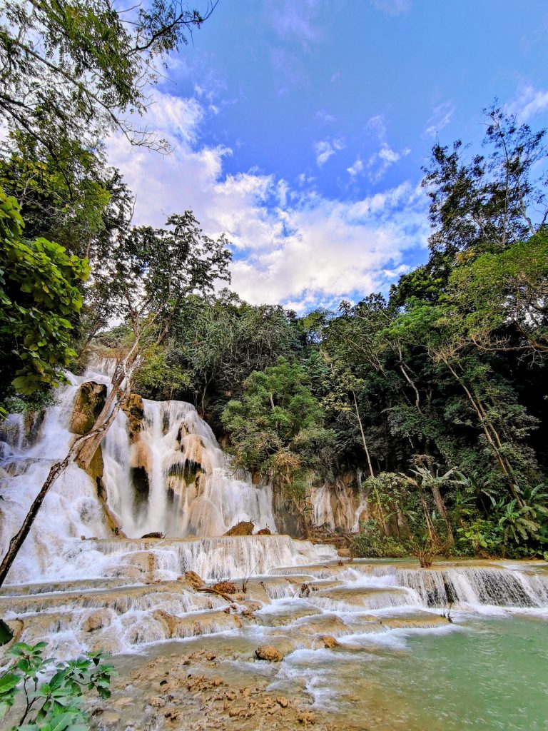 Kuang Si Waterfalls near Luang Prabang, Laos, flow gently over layered limestone terraces into clear turquoise pools, framed by lush jungle and tall trees, offering a peaceful side view of one of the country’s most beautiful natural attractions.