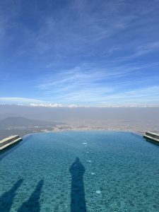 A stunning infinity pool extends towards a picturesque landscape, reflecting the clear blue sky above. Snow-capped mountains are visible in the distance, surrounded by a hazy view of a valley below, while the pool's surface features shimmering tiles. 