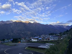 A small colorful town with a curved runway in the foreground, set below dramatic snow-capped mountains under a partly cloudy sky.