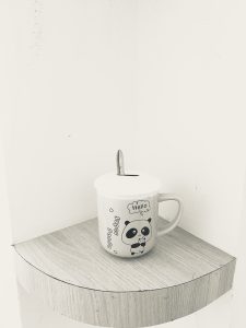 A black and white photo of a decorative mug with a lid and spoon resting on a light-colored wooden surface.