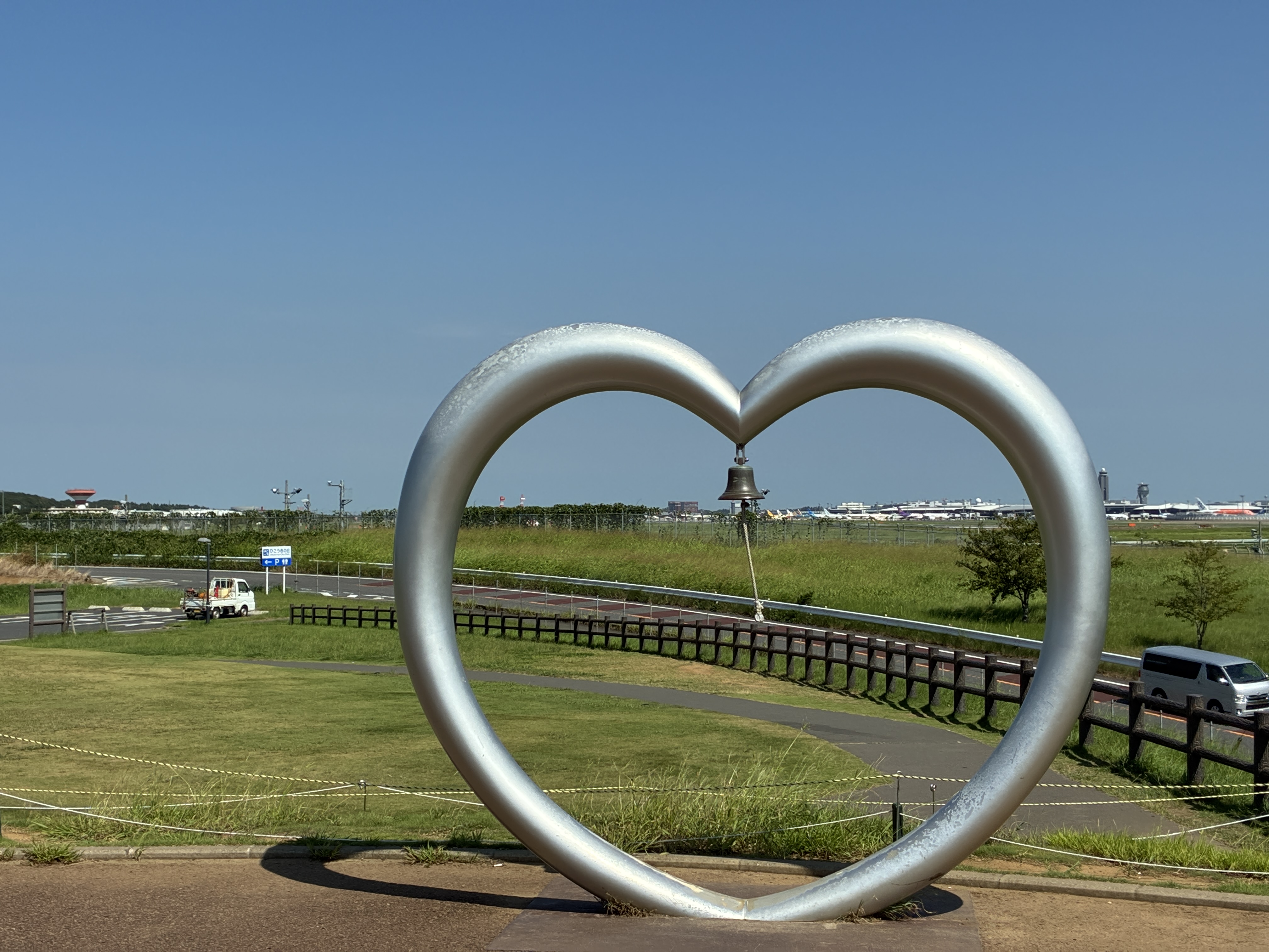 A large metallic heart sculpture stands prominently in the foreground, with a bell hanging in its center