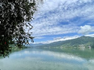 Still water of Phewa Lake mirrors clouds and distant hills, framed by trees in Pokhara, Nepal, capturing a serene and refreshing moment. 