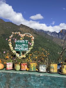 A heart-shaped floral sign reading “EVEREST ROUTE GOOD LUCK,” surrounded by colorful flower pots, with green hills and mountains under a blue sky.