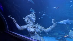 A stone statue of a female mythical figure with arms raised submerged in a blue aquarium tank with fish.