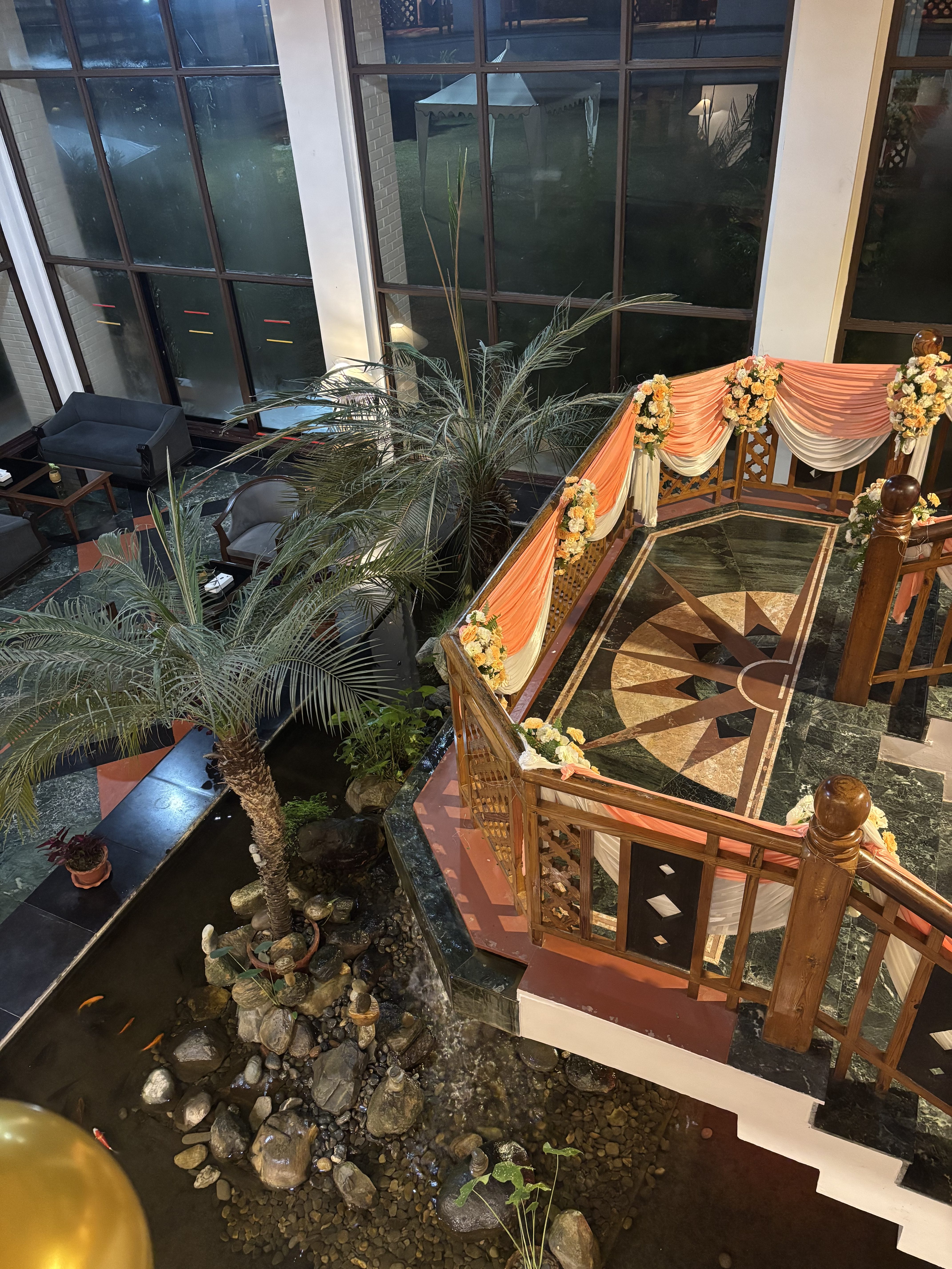 A spacious indoor area featuring modern furnishings and large windows, with a decorative railing adorned with orange and white drapes and floral arrangements. Below, a small pond with rocks and aquatic plants houses colorful koi fish.