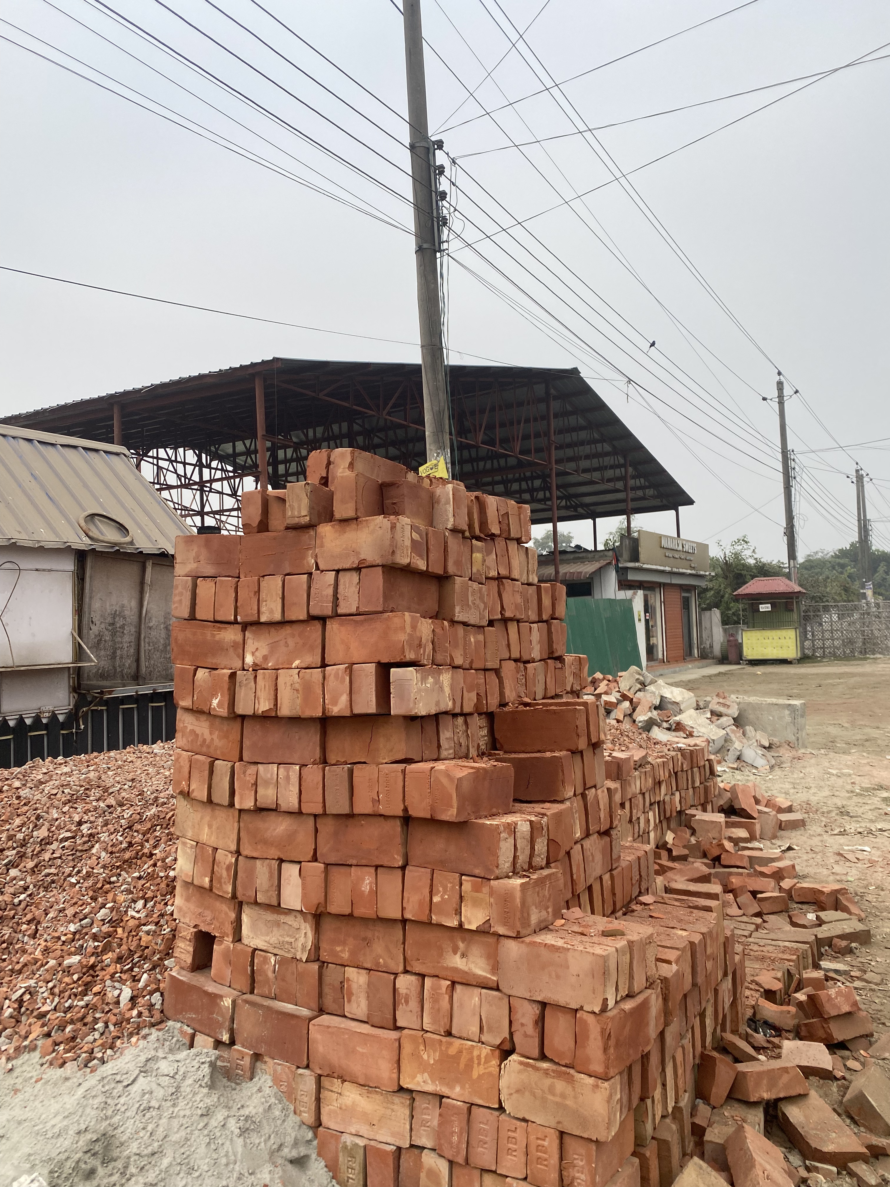 Stacked red bricks sit beside a dirt road under overcast skies. Nearby, a metal-roofed structure and power lines emphasize an industrial setting.