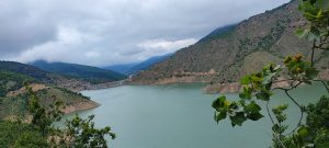 A vivid emerald landscape surrounds the calm blue waters of Suleiman Tangeh Dam Lake in Mazandaran, northern Iran, with dense green forests covering the hillsides