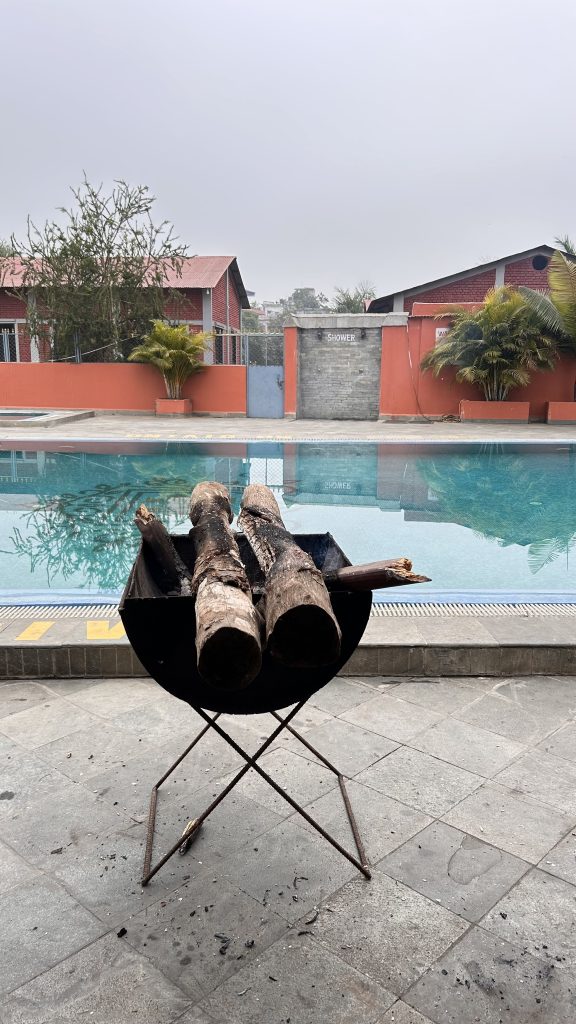 A black fire pit with several logs of wood placed on top sits on a tiled surface near a blue swimming pool.