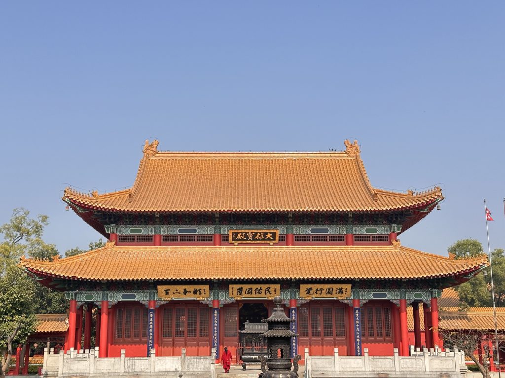 A traditional Chinese-style Lumbini temple with a curved tile roof, decorated in red and gold with detailed architectural designs.