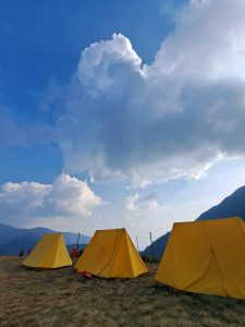 A scenic view of several yellow tents set up on a grassy plain, with a backdrop of mountains and a bright blue sky filled with fluffy clouds. 