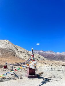 A rocky, barren landscape with a red stupa wrapped in colorful prayer flags under a clear blue sky.