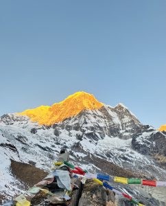 A panoramic view of a snow-capped mountain peak illuminated by the golden light of sunrise. In the foreground, colorful prayer flags are strewn across rocky terrain, contrasting with the stark white of the snow and the earthy brown of the ground. 