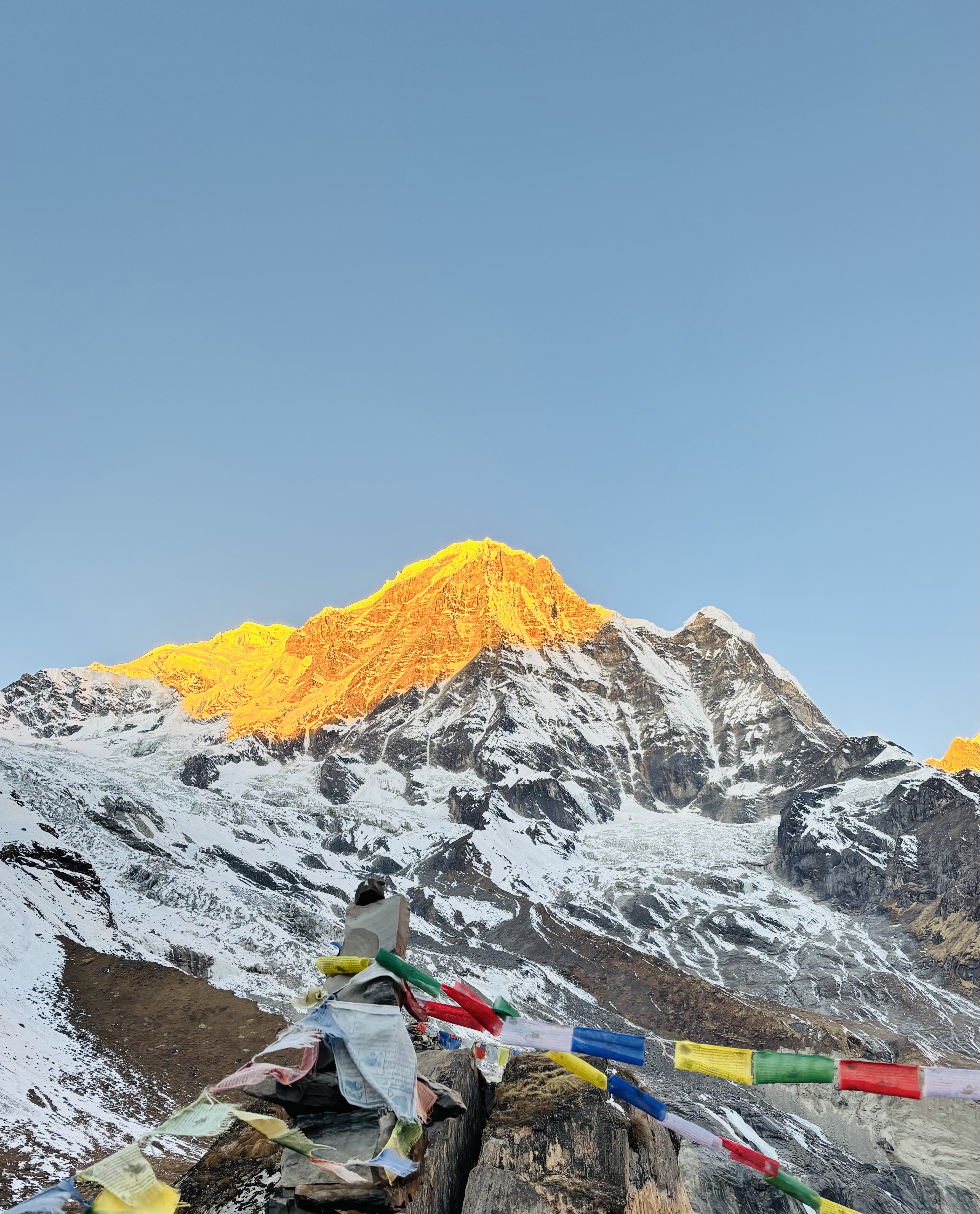 A panoramic view of a snow-capped mountain peak illuminated by the golden light of sunrise. In the foreground, colorful prayer flags are strewn across rocky terrain, contrasting with the stark white of the snow and the earthy brown of the ground.