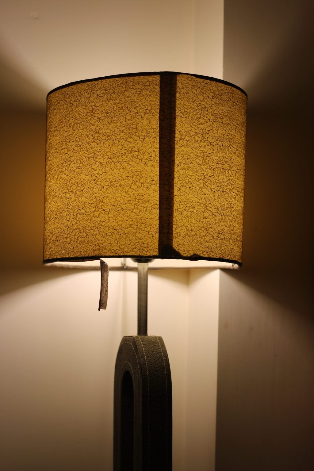 Decorative table lamp illuminated. Lamp shade has an erratic wiggly line design, the maker’s seem fabric join is clearly visible. An edge binding tape hangs in need of repair.
