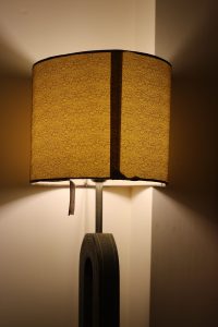 Decorative table lamp illuminated. Lamp shade has an erratic wiggly line design, the maker’s seem fabric join is clearly visible. An edge binding tape hangs in need of repair.
