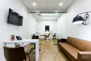 A modern, warm waiting room featuring a glass partition, brown leather seating, wooden floors, and minimal décor including a small table with a phone and photo, a wall-mounted TV, stylish lighting, and cat artwork. 