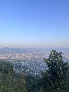 A panoramic view of a sprawling city nestled in a valley, surrounded by distant mountains under a clear blue sky. 