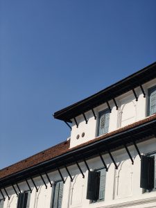A close-up view of a building's upper facade featuring white walls with decorative elements, a sloped red-tiled roof, and multiple blue wooden shutters that are partially open.
