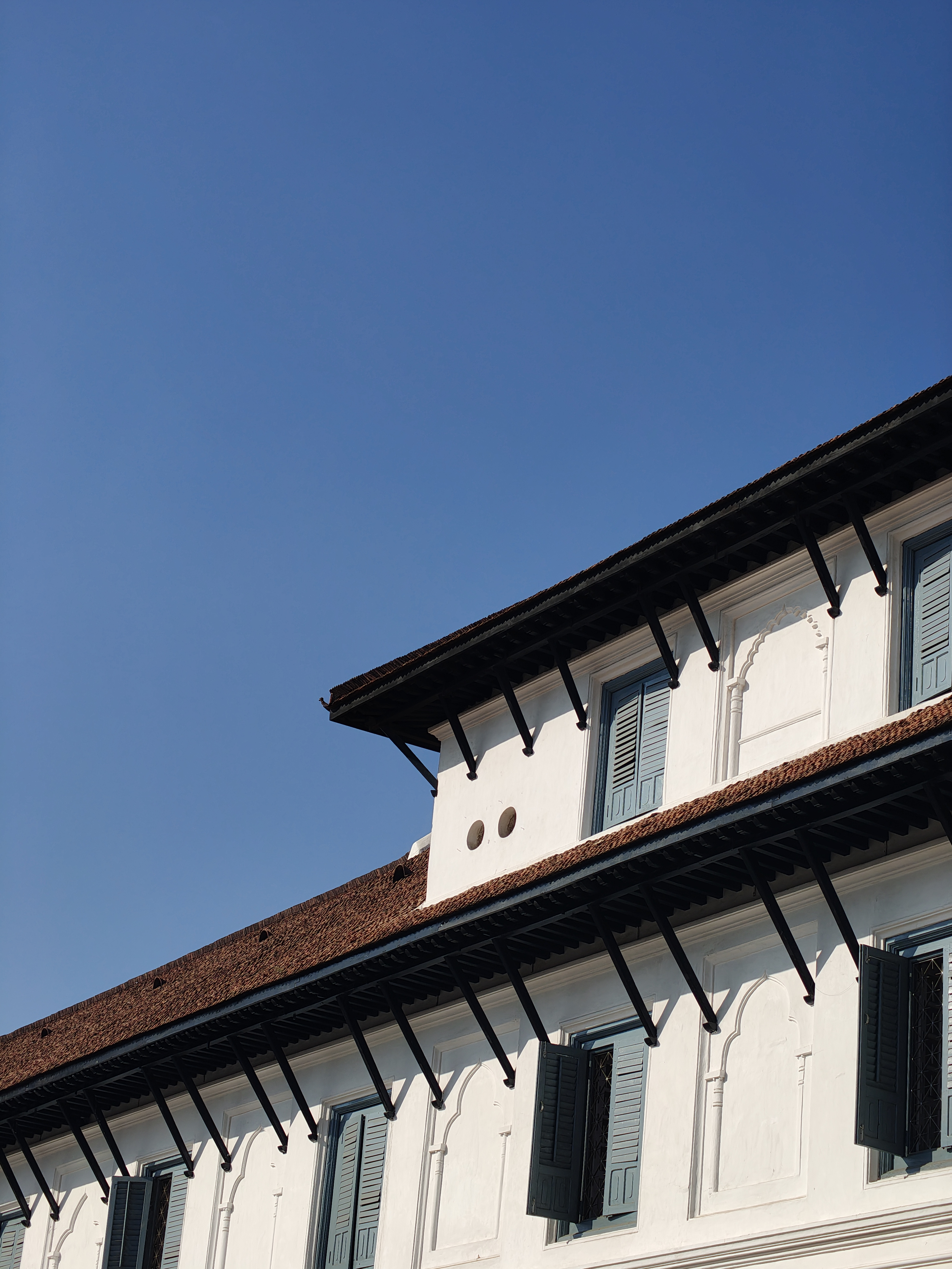 A close-up view of a building's upper facade featuring white walls with decorative elements, a sloped red-tiled roof, and multiple blue wooden shutters that are partially open.