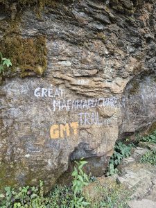 A rocky surface with green moss and plants at the base, featuring painted text reading “GREAT MACCHAPUCHRE TRAIL” in white and “GMT” in orange.