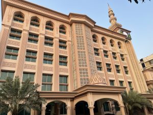 A low-angle shot of a multi-story peach-colored Islamic building featuring traditional arched windows and intricate geometric latticework. A tall, slender minaret with a digital clock face rises into a clear blue sky. Palm trees are visible at the base of the structure near an arched entryway.
