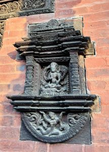 
A detailed wooden relief depicting a deity seated in a meditative pose surrounded by intricate floral and geometric designs. Above the deity, there are ornate, carved architectural elements.