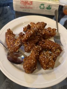 A plate of crispy, golden-brown chicken wings garnished with sesame seeds on a table. They appear glazed and shiny, with a spoon and fork beside.