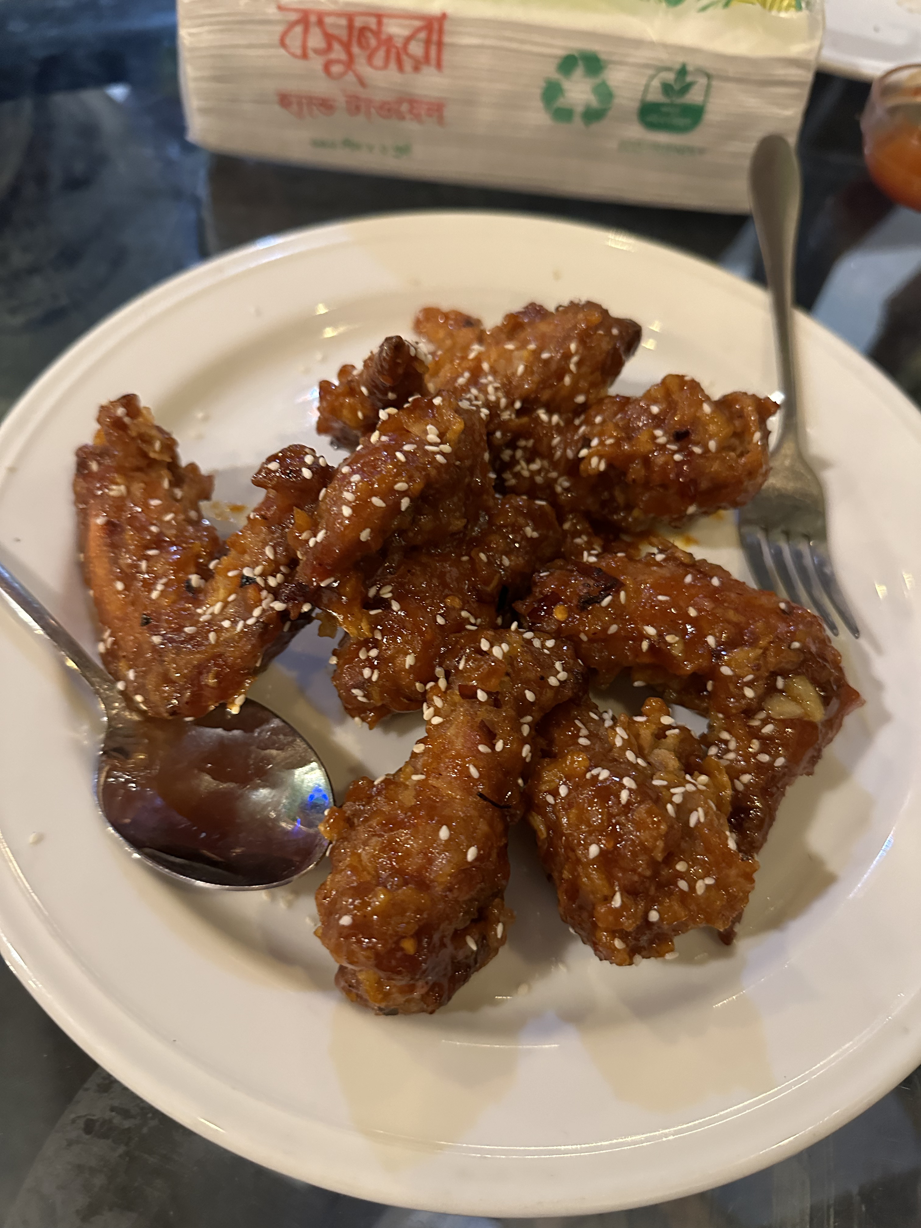 A plate of crispy, golden-brown chicken wings garnished with sesame seeds on a table. They appear glazed and shiny, with a spoon and fork beside.