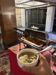 A hand holds a small white bowl containing clear broth with pieces of chicken and green vegetables. In the background, there is a buffet setup with a silver chafing dish holding a dish that appears to contain a sauce and pieces of food
