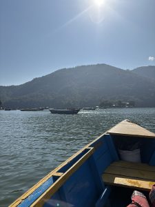 
A view from inside a small wooden boat with a blue and yellow color scheme, looking out over calm water towards a mountainous landscape under a clear blue sky.