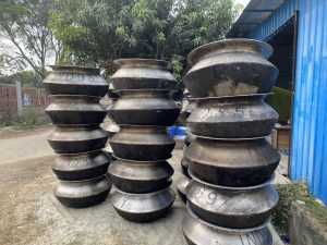 Tall stacks of large, metallic, industrial pots are arranged outdoors beside a blue corrugated shed. Green trees and a brick wall are in the background.