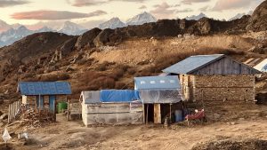 A small group of buildings in a rugged mountain area, with simple structures in the foreground and snow-capped mountains beneath a pink and blue sky.