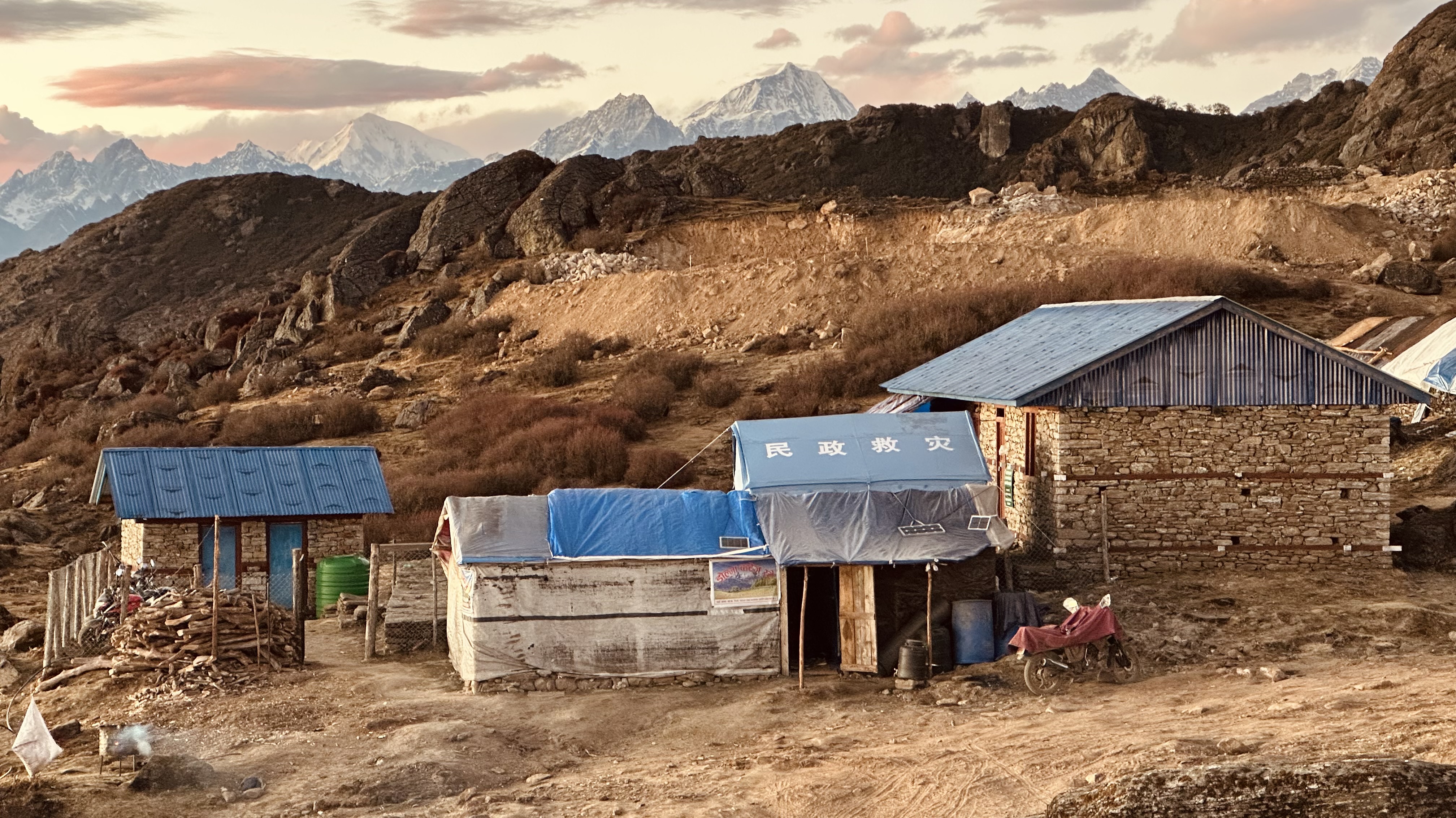 A small group of buildings in a rugged mountain area, with simple structures in the foreground and snow-capped mountains beneath a pink and blue sky.