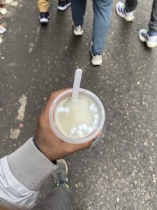 A close-up of a hand holding a cup of coconut water with ice and a straw on a busy street with people walking in the background.