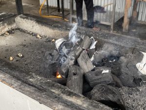 A close-up view of a charcoal grill with burning charcoal, producing smoke and a small flame.