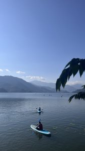 Two people paddleboarding on a calm lake under a clear blue sky, surrounded by mountains in the background.