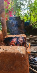 A close-up of mutton barbecue grilling on metal skewers over an open flame on a rustic stone and dirt grill.