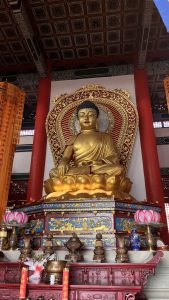 A golden Buddha statue inside a Chinese Buddhist temple in Lumbini, seated on a lotus throne. Red pillars and hanging prayer banners surround it, creating a calm and sacred atmosphere. The walls of the temple are decorated with colorful patterns and lanterns, while various ornamental objects and offerings, including vases and candles, are placed on a low wooden altar in front of the statue. The ceiling features ornate designs and is highlighted by beams painted in vibrant colors.