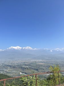 A panoramic view of mountains with snow-capped peaks against a clear blue sky, overlooking a sprawling valley and city below. 