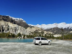 A white SUV is parked on rocky ground beside a blue lake in Manang, Nepal. Tall mountains rise in the background, some topped with snow, contrasting with the deep blue sky.

