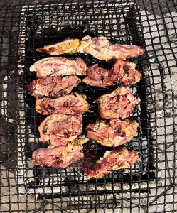A grilling surface with several pieces of marinated meat is visible, sizzling over hot coals.